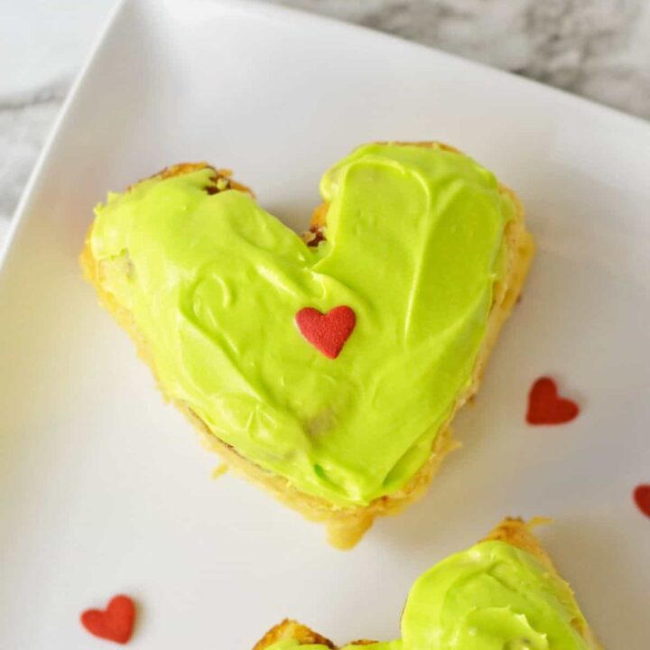 Two heart shaped grinch cinnamon rolls with green frosting and heart sprinkles.