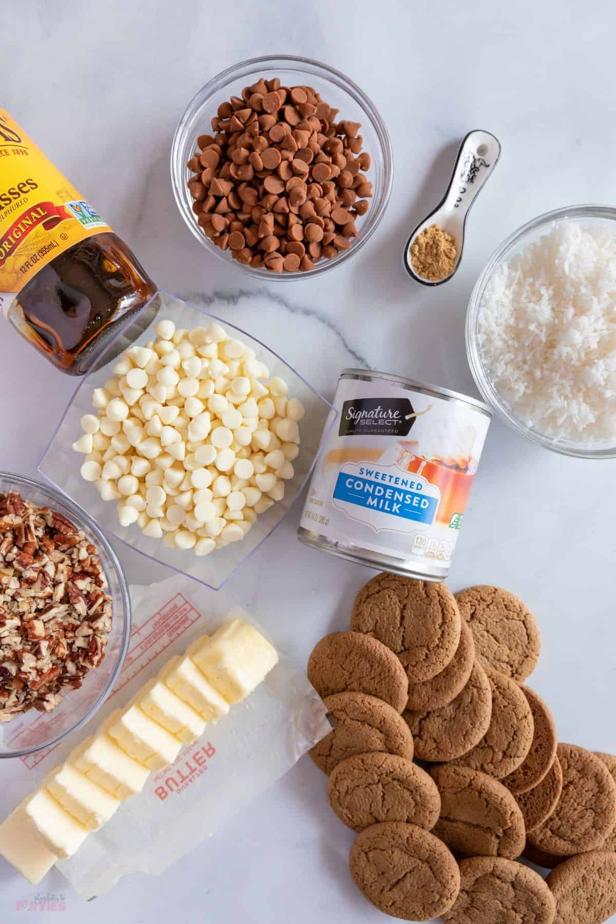 Ingredients on a marble surface include molasses, white chocolate chips, cinnamon chips, ginger, condensed milk, coconut, butter, pecans, and gingersnaps.