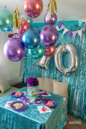A corner of a room decorated for a 10th birthday party including a balloon chandelier, fringe curtain, and crushed velvet tablecloth.