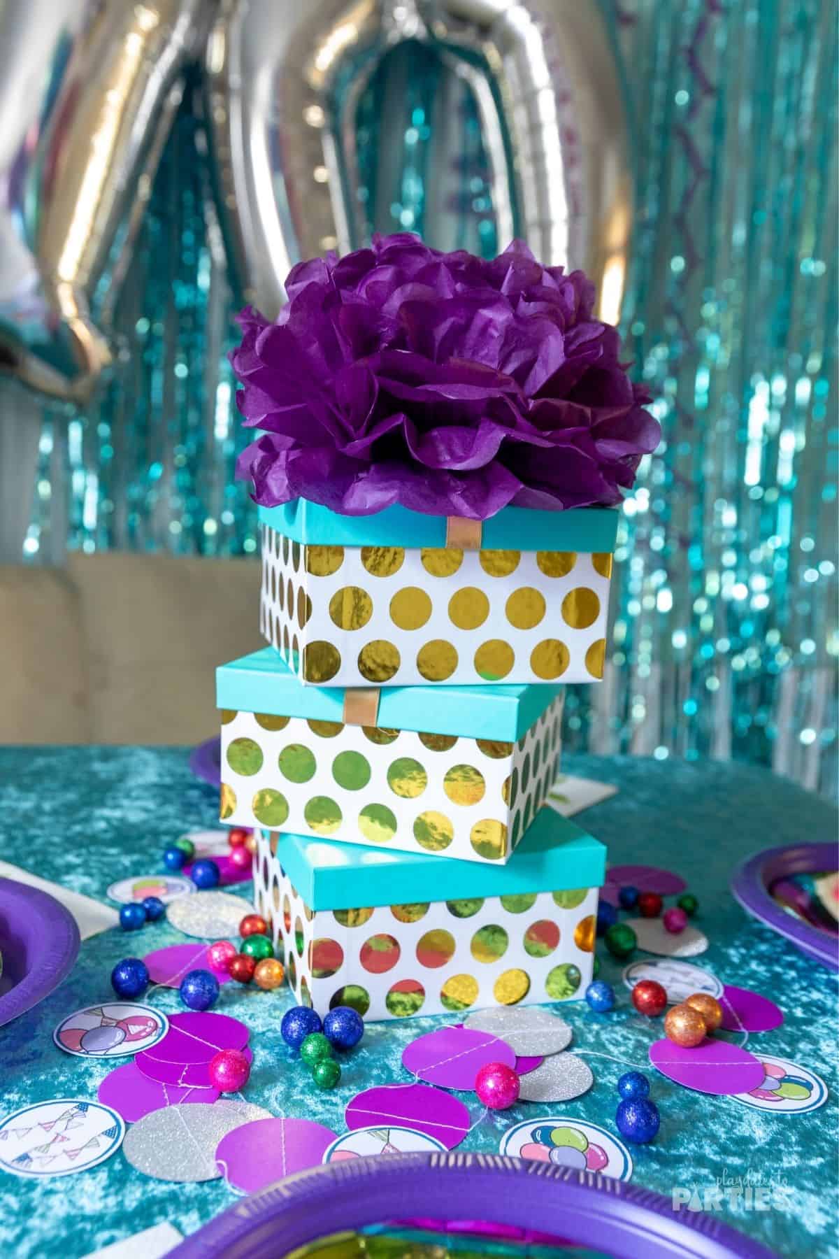 A stack of gold polka dot party favor boxes with blue lids and purple decor around them.