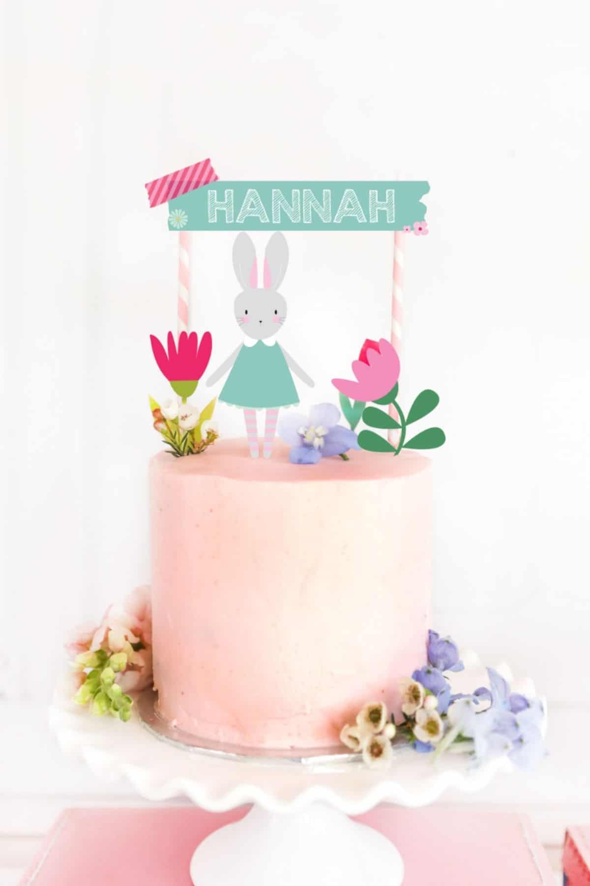 A pastel pink birthday cake with a spring topper including a bunny, flowers, and the birthday girls name.