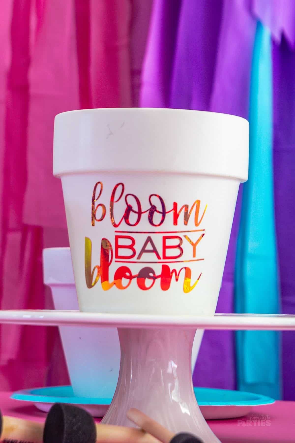 A white pot on a cake stand that says bloom baby bloom in hologram letters.