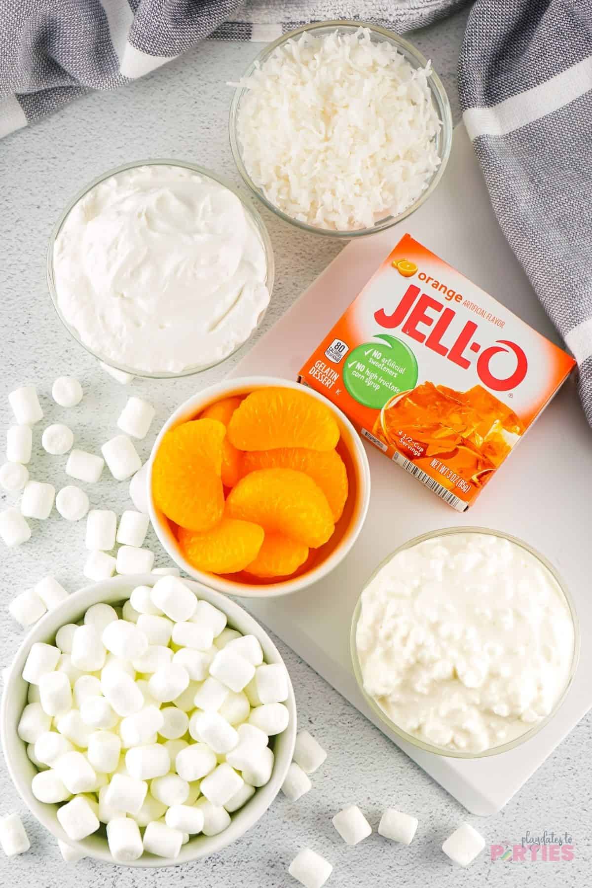 Ingredients on a table including orange jello, mandarins, marshmallows, coconut, cool whip, and cottage cheese.