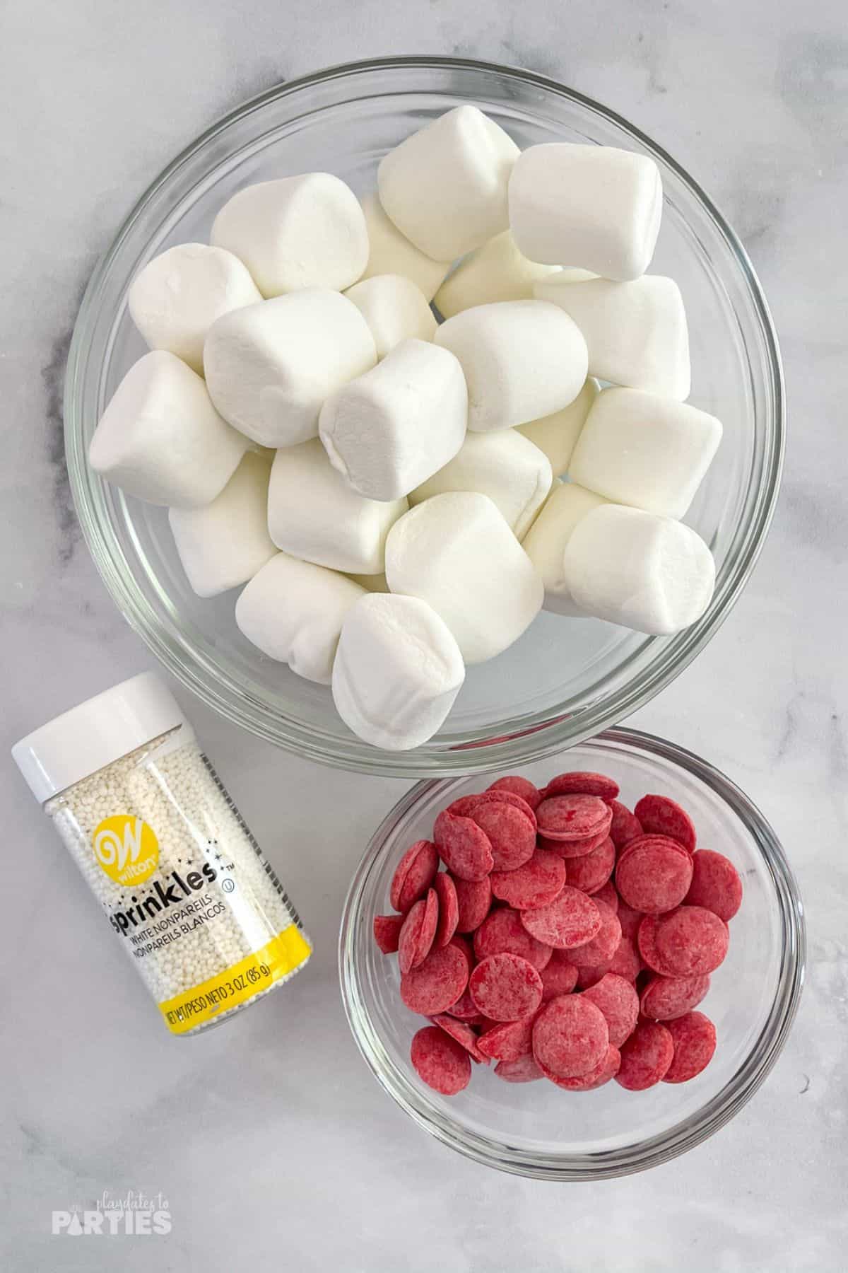 Ingredients on a white surface - marshmallows, red candy melts, and white sprinkles.