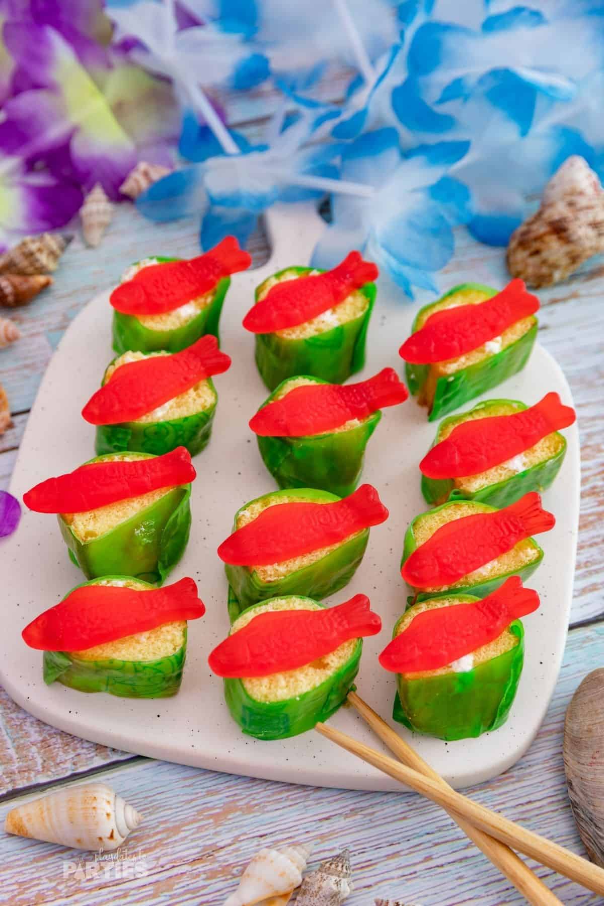Candy sushi on a white cutting board with luau decorations nearby.