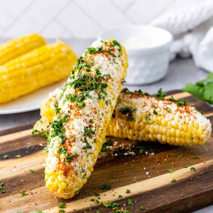 Two mexican elotes corn cobs on a cutting board.