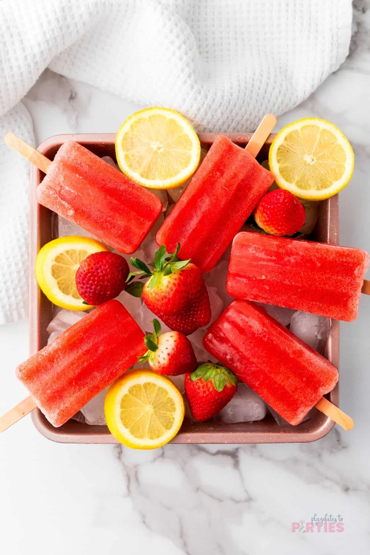 Homemade strawberry popsicles are nestled on a bowl of ice cubes with fresh strawberries and lemon slices.