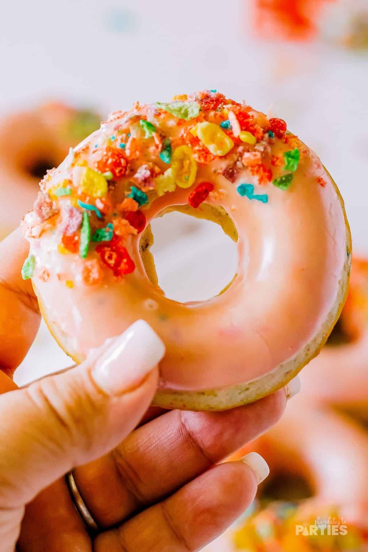 A woman's hand holding a baked donut with strawberry glaze and colorful cereal.