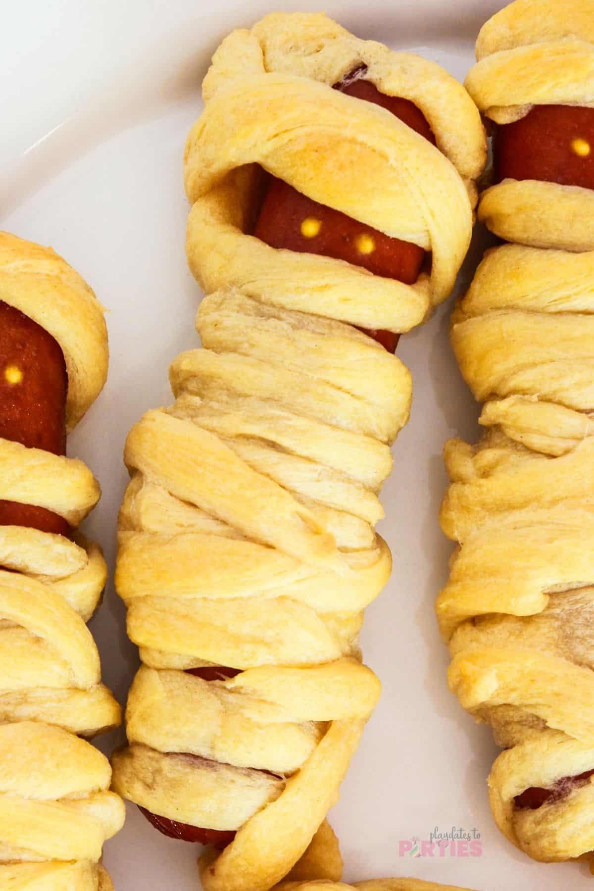Crescent roll mummy hot dogs on a white plate.