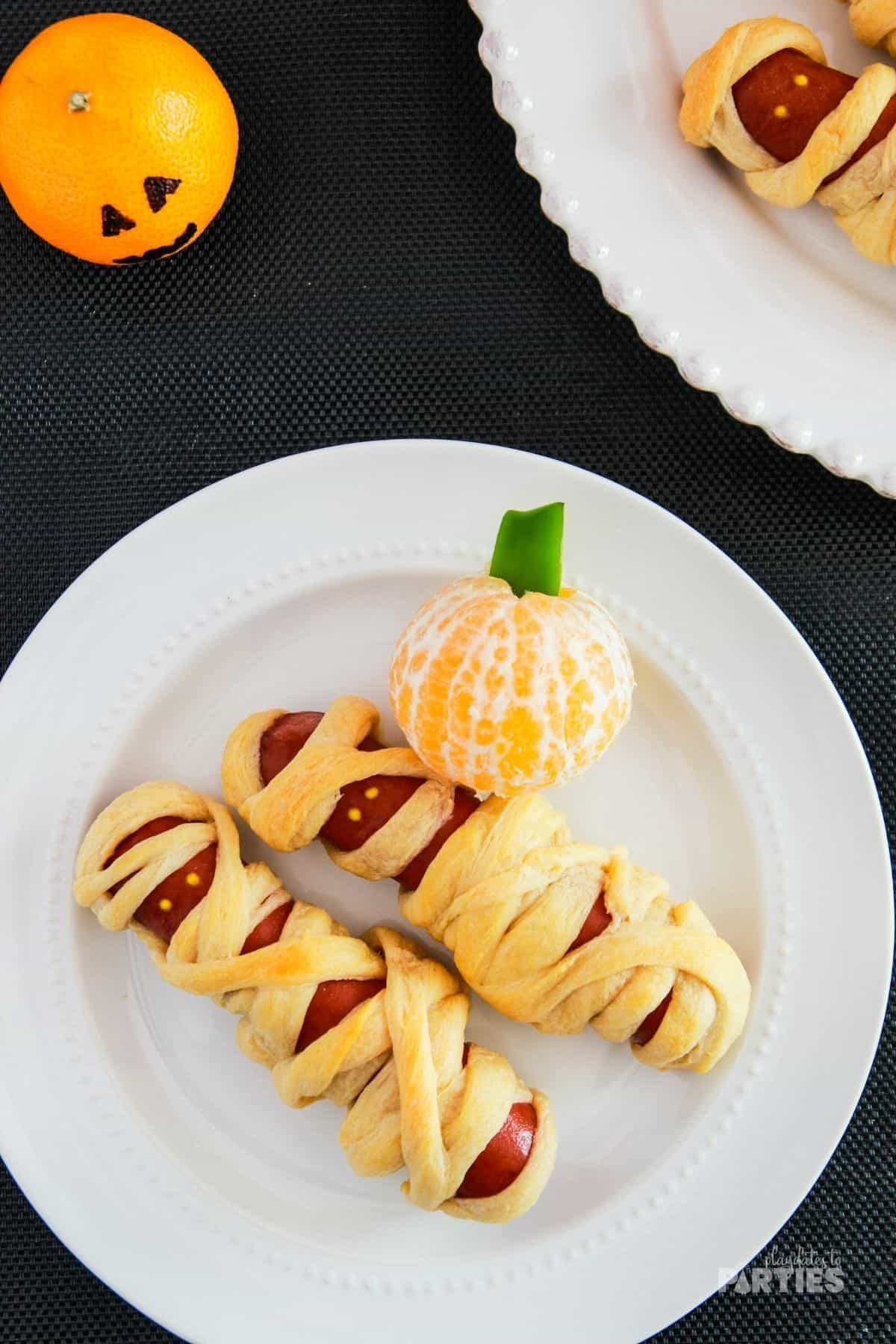 Mummy hot dogs on a plate with a clementine pumpkin.