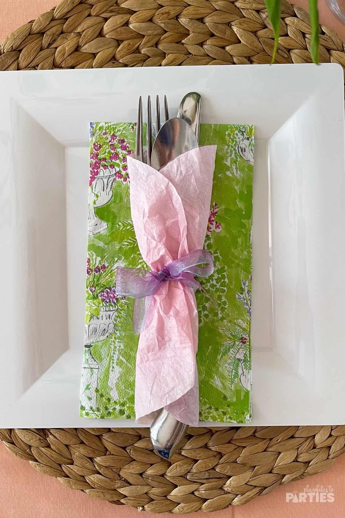 Utensils in a pink wrapper layered on top of a green napkin and a white plate.