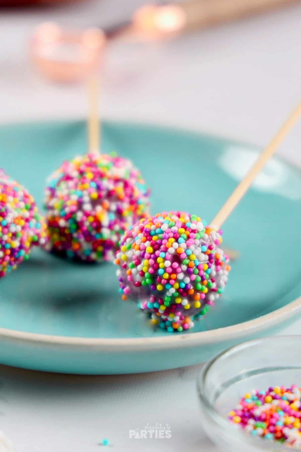 Bite sized cake ball on a stick covered in rainbow sprinkles.