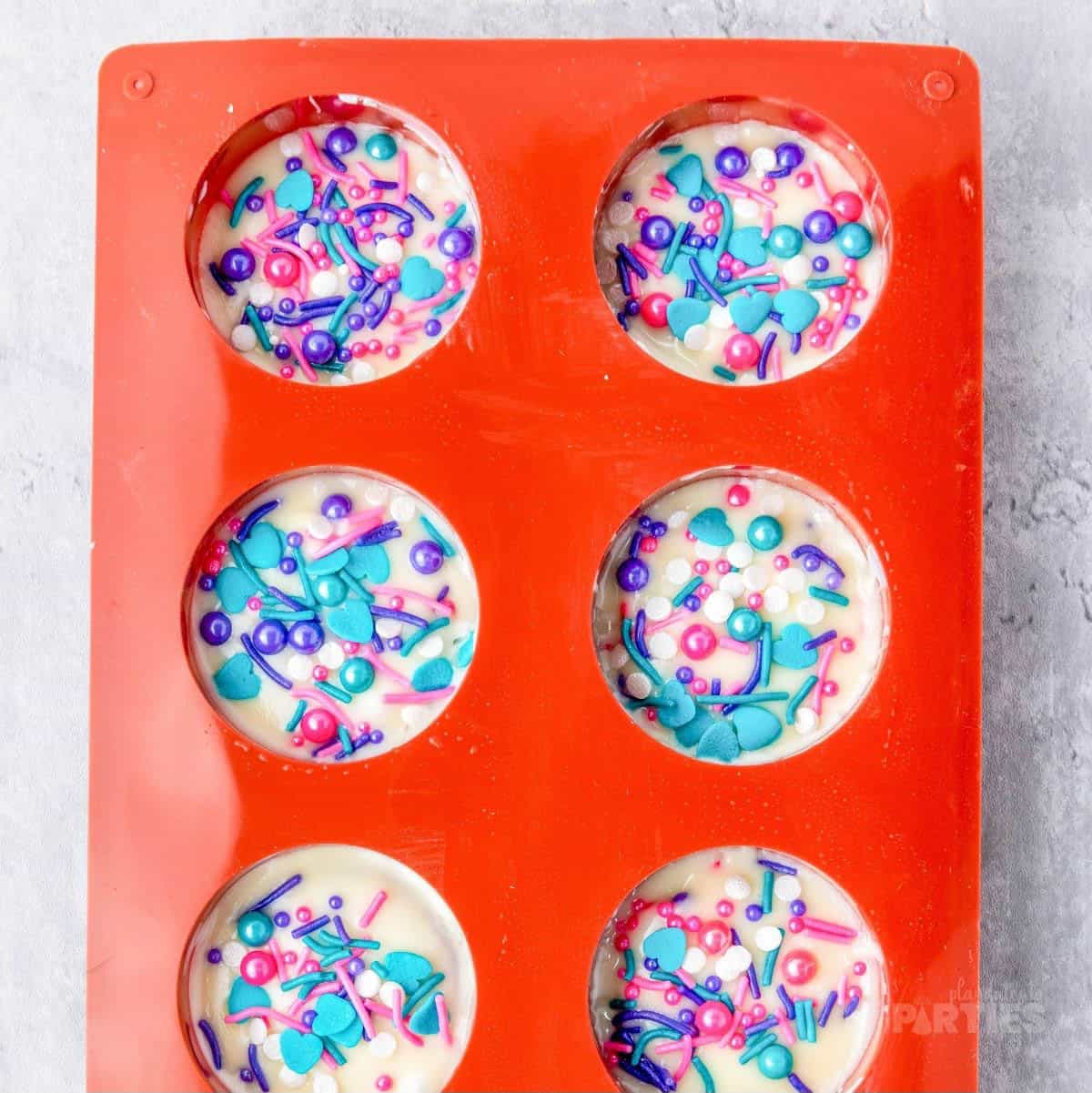 A mold filled with white chocolate and decorated with purple, blue, and pink sprinkles.