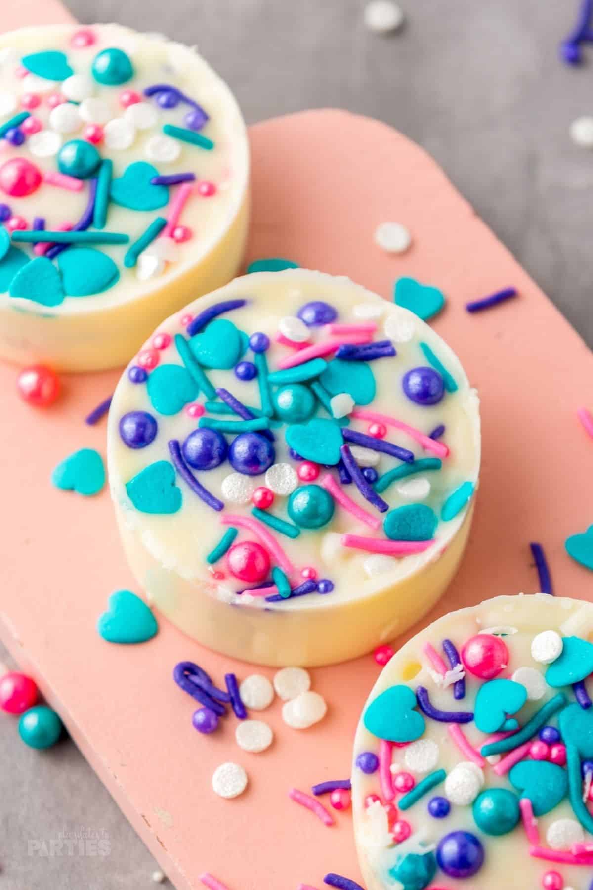 Three white chocolate covered Oreos covered in sprinkles sit on a pink board