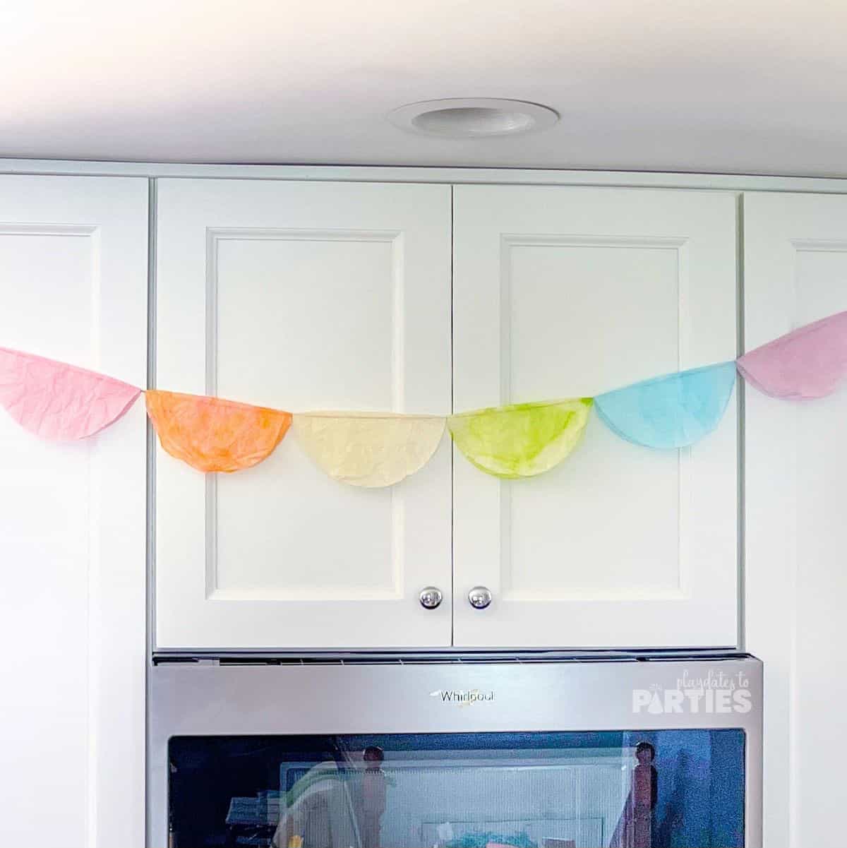 A rainbow colored coffee filter banner hung across white kitchen cabinets.