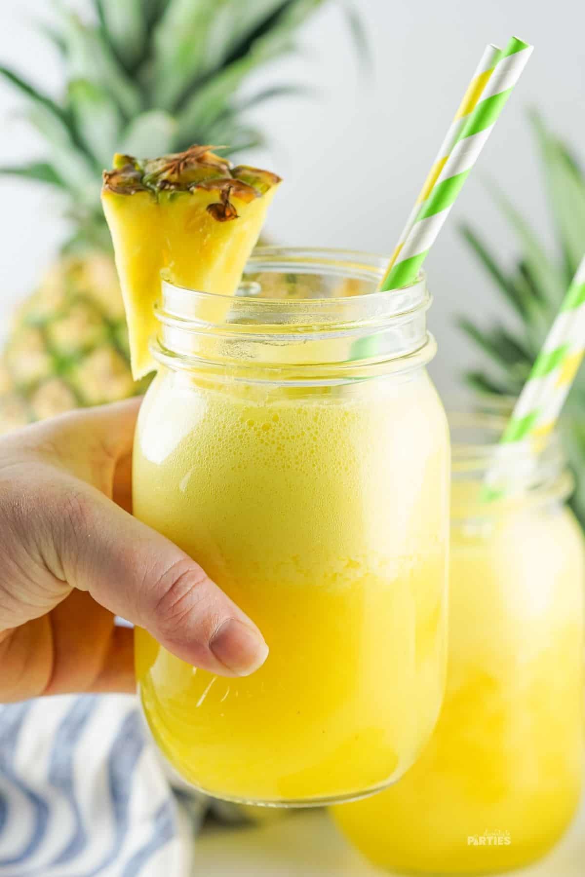 A woman's hand holds a glass filled with a frothy yellow beverage garnished with a pineapple.