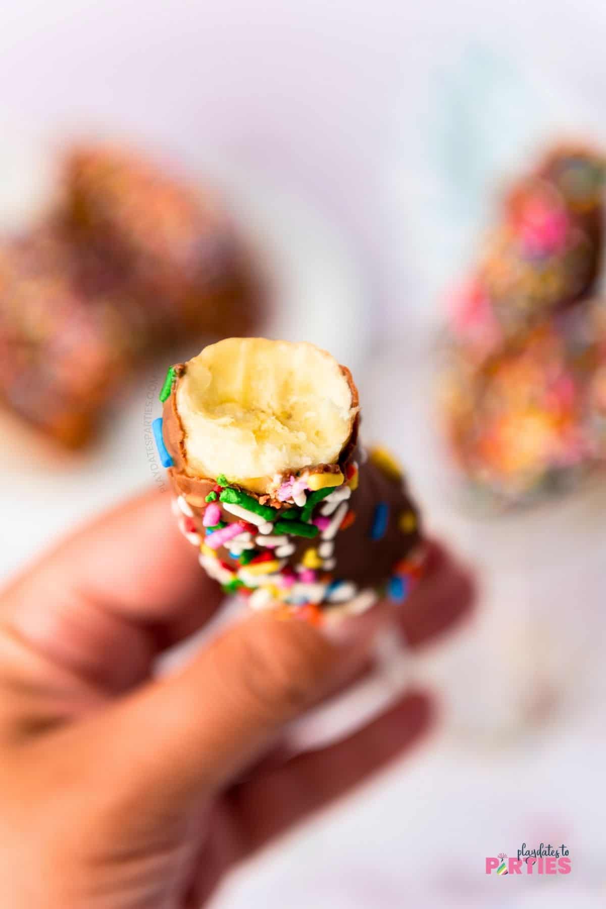 A woman's hand holding a chocolate covered banana with a bite taken out.