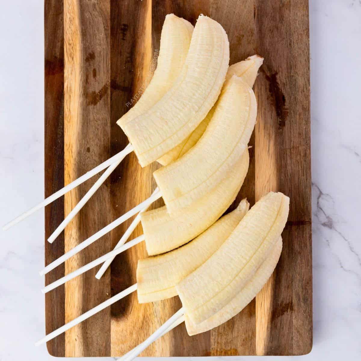 Several banana halves with lollipop sticks on a cutting board.