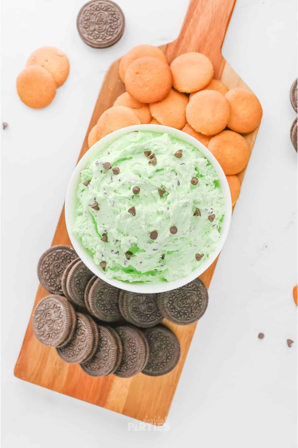 A wood board with cookies and a bowl of mint chocolate chip dip.